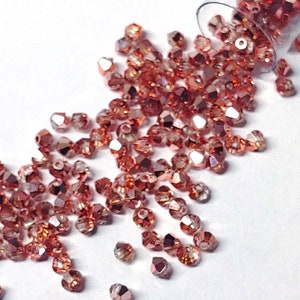 May include: A close-up of a pile of small, rose gold-colored glass beads. The beads are faceted and have a shimmery finish.