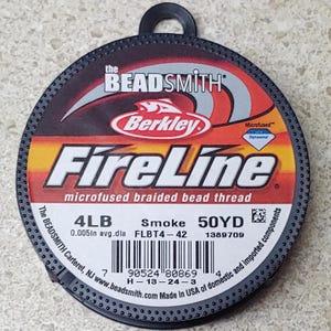 May include: A spool of black FireLine microfused braided bead thread by Berkley. The spool is labeled with the text "4LB 0.005in avg.dla Smoke FLBT4-42 50YD 1389709".