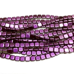 May include: Close-up of strands of square-shaped beads in a deep purple hue. The beads have a metallic sheen, reflecting light. The beads are strung together, creating a textured surface. The beads are likely for jewelry making.