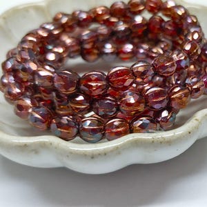 May include: A strand of iridescent red glass beads with a faceted, oval shape. The beads are arranged in a coil on a white ceramic dish.