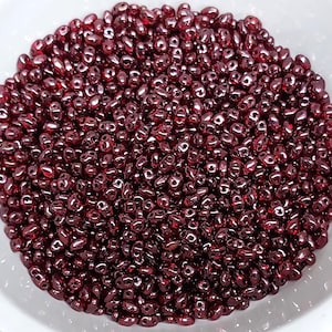 May include: A bowl filled with small, dark red glass beads. The beads are oval shaped and have a glossy finish.