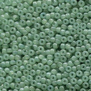May include: A close-up view of a pile of small, round, translucent beads in a pale green color. The beads have a hole through the center and are densely packed together, creating a textured surface.