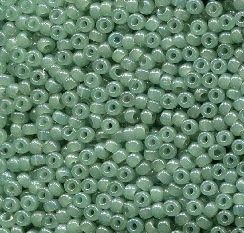 May include: A close-up view of a pile of small, round, translucent beads in a pale green color. The beads have a hole through the center and are densely packed together, creating a textured surface.