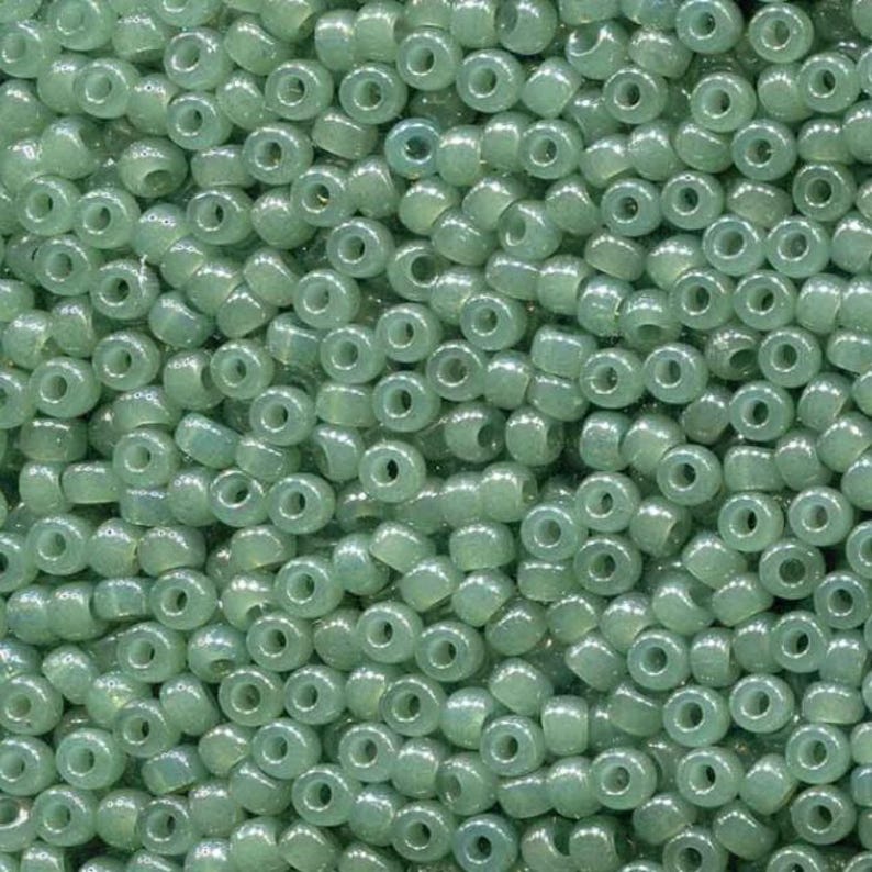 May include: Close-up of a pile of small, round, translucent beads in a pale green color. The beads have a hole through the center and are densely packed together, creating a textured surface. Ideal for jewelry making and crafts.