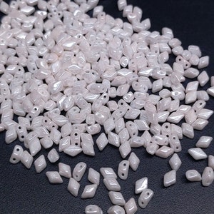 May include: A pile of white iridescent glass beads in a diamond shape. The beads have two holes in each bead.