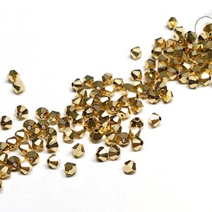 May include: A close-up of a pile of gold-colored glass beads. The beads are faceted and have a shiny finish. They are scattered on a white surface.