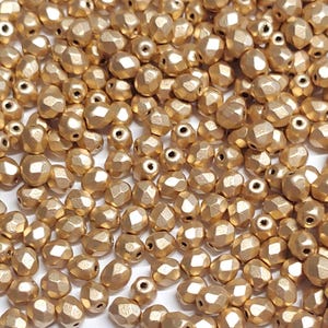 May include: A close-up of a pile of gold-colored faceted glass beads. The beads are small and have a matte finish.