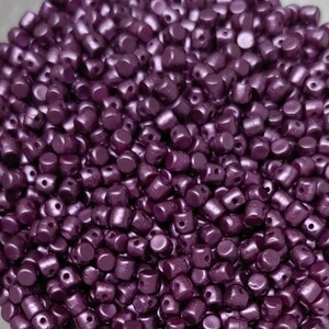 May include: A close-up view of a large quantity of small, round, purple beads with a metallic sheen. The beads have two holes in each one.