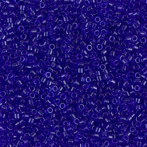 May include: A close-up shot of numerous small, cylindrical, transparent, dark blue glass beads. The beads are scattered, creating a dense, textured surface. These beads are commonly used for jewelry making and crafting.