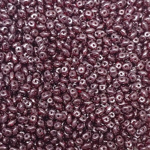 May include: A close-up shot of a pile of dark red, two-holed beads. The beads are small, with a glossy finish, and appear to be made of glass or a similar material. The beads are scattered, creating a textured surface.