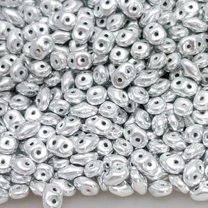 May include: A close-up of a collection of silver beads. Each bead has an irregular shape with two holes. The beads have a metallic finish, reflecting light. The beads are scattered, creating a textured surface. The beads are suitable for jewellery making.