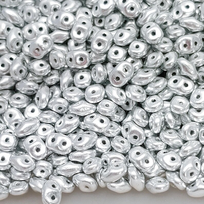 May include: A close-up of a collection of silver beads. Each bead has an irregular shape with two holes. The beads have a metallic finish, reflecting light. The beads are scattered, creating a textured surface. The beads are suitable for jewellery making.