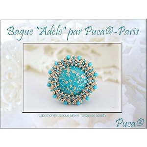 May include: A turquoise ring with a gold beaded border. The ring features a turquoise cabochon with a gold flecked design. The ring is labeled "Bague "Adèle" par Pucca® - Paris" and "Cabochon® Opaque Green Turquoise Splash".