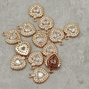 May include: A group of gold-tone heart-shaped charms with clear crystal embellishments. Each charm has a loop at the top for attaching to jewelry or other crafts.