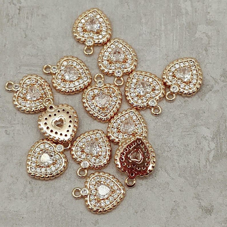 May include: A group of gold-tone heart-shaped charms with clear crystal embellishments. Each charm has a loop at the top for attaching to jewelry or other crafts.