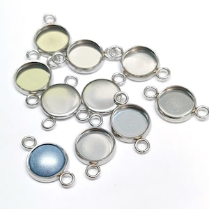 May include: A group of silver-toned metal bezel settings with three loops for attaching to jewelry. The bezels are round and have a clear glass insert. The bezels are arranged in a random pattern on a white surface.