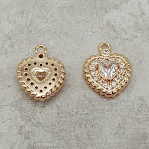 May include: Two gold heart-shaped charms with a decorative border. One charm has a clear rhinestone in the center, while the other has a smaller heart-shaped cutout in the center.