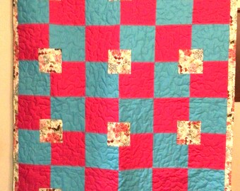 May include: A handmade quilt with a checkerboard pattern of bright pink and turquoise squares. Each turquoise square has a small floral square in the centre. The quilt is trimmed with a floral border and hung on a wooden rod.