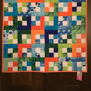 Könnte beinhalten: Ein farbenfrohes Patchwork-Quilt mit einer Mischung aus orangefarbenen, blauen, grünen und weißen Quadraten. Das Quilt weist ein sich wiederholendes Muster aus Quadraten und Rechtecken mit einem kontrastierenden orangefarbenen Rand auf. Das Quilt hängt an einer dunklen Holztür.