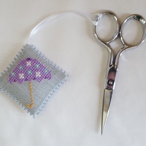 May include: A cross-stitched square featuring a purple umbrella design, edged with blue beads, and a white ribbon. Next to it, a pair of small, silver scissors.