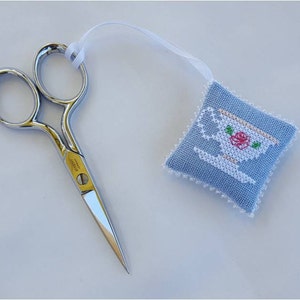 Puede incluir: Un pequeño protector de agujas azul y blanco con un diseño de taza de té blanca y una rosa rosa. El protector de agujas está unido a una cinta blanca y un par de tijeras plateadas.