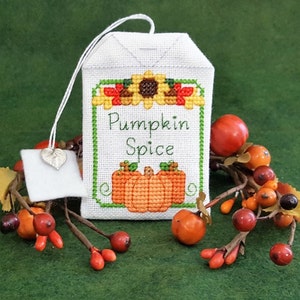 May include: A white tea bag with a cross-stitch design featuring a sunflower and pumpkins. The tea bag is labeled "Pumpkin Spice".
