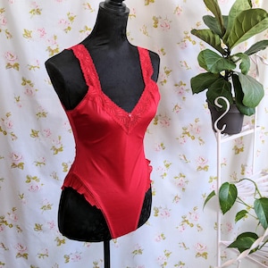 May include: Red satin bodysuit with lace trim and ruffled detailing. The bodysuit is on a black mannequin.