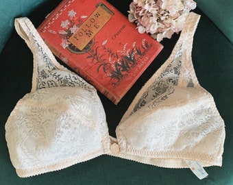 May include: White lace bra with a delicate floral pattern. The bra has adjustable straps and a hook and eye closure. The bra is lying on a green surface with a book titled "Follow Me" on top of it.
