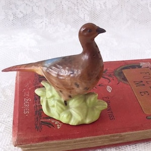 May include: A small ceramic figurine of a brown and white pheasant standing on a green base. The figurine is sitting on top of a red book with the title "Follow Me" on the cover.