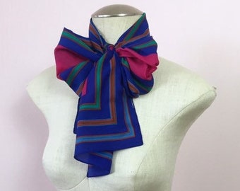 May include: A blue and pink striped scarf tied in a bow on a white mannequin. The scarf has a geometric pattern with green and brown stripes.