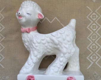 May include: White ceramic lamb figurine with pink accents, riding on a white platform with pink wheels. The lamb has a pink bow around its neck and closed eyes.
