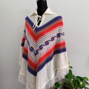 May include: White knitted poncho with a geometric pattern in red, blue, and orange. The poncho has a fringe trim and a tie at the neck.