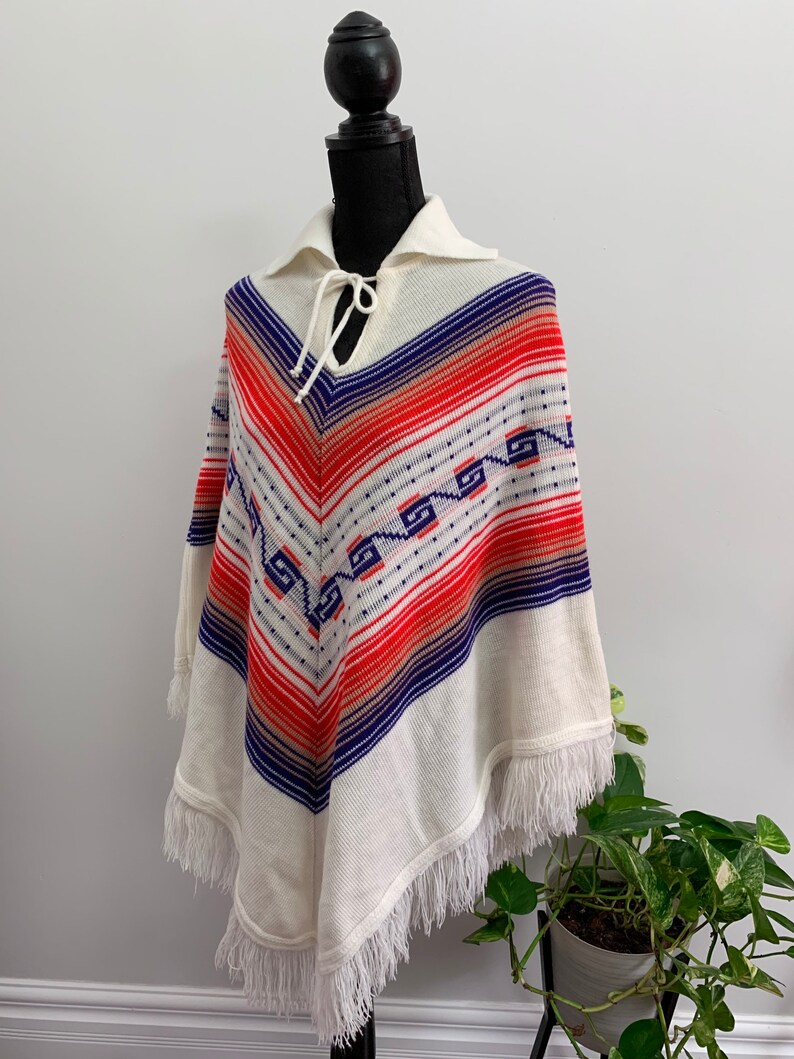 May include: White knitted poncho with a geometric pattern in red, blue, and orange. The poncho has a fringe trim and a tie at the neck.