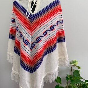 May include: White knitted poncho with a blue, red, and orange geometric pattern. The poncho has a collar and a tie at the neck. The poncho has fringe around the edges.
