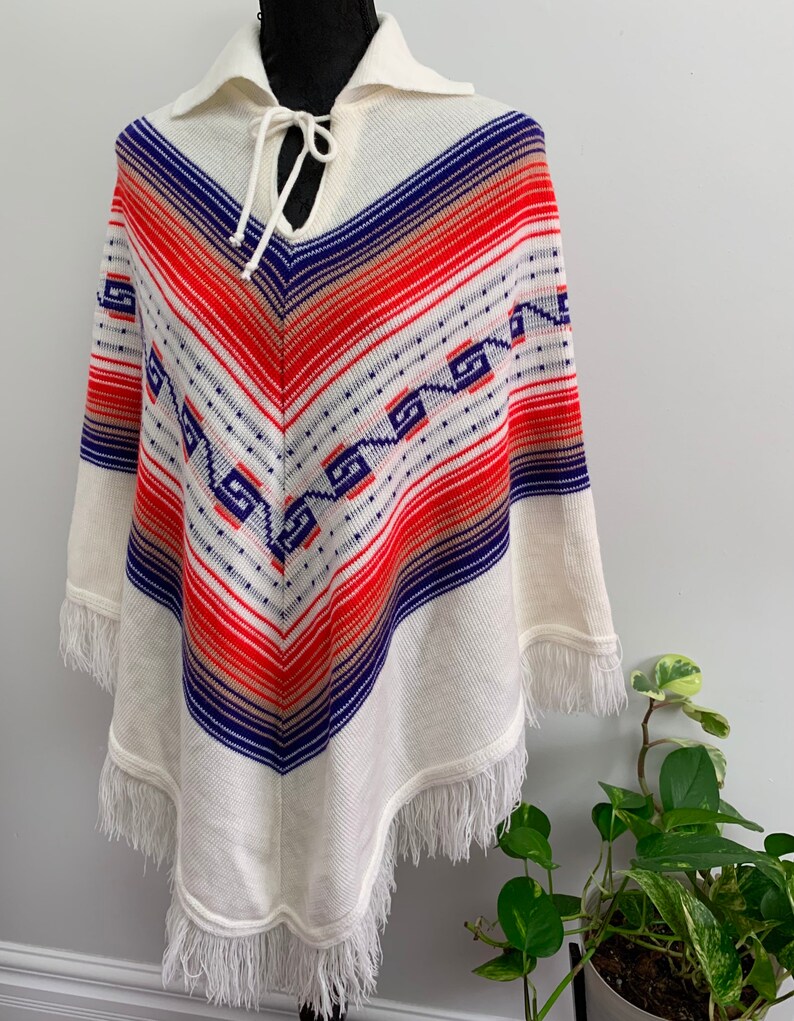 May include: White knitted poncho with a blue, red, and orange geometric pattern. The poncho has a collar and a tie at the neck. The poncho has fringe around the edges.