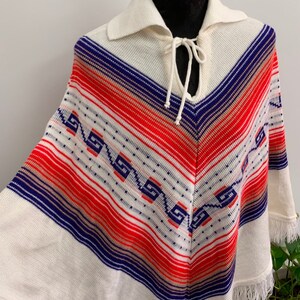 May include: White knitted poncho with a blue, red, and beige striped pattern and a fringe trim. The poncho has a collar and a tie at the neck.