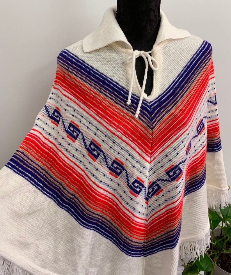 May include: White knitted poncho with a blue, red, and beige striped pattern and a fringe trim. The poncho has a collar and a tie at the neck.