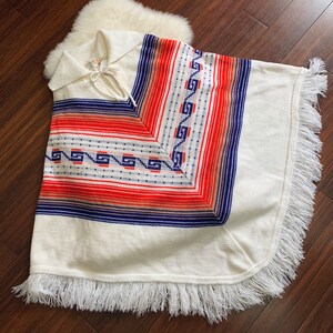 May include: White knitted poncho with a blue, red, and white geometric pattern. The poncho has a fringe border and a tie at the neck.