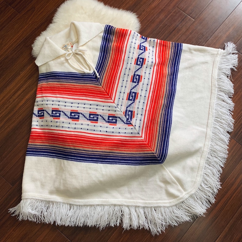 May include: White knitted poncho with a blue, red, and white geometric pattern. The poncho has a fringe border and a tie at the neck.