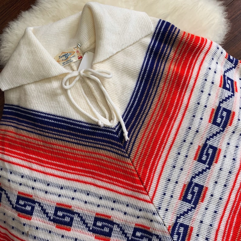 May include: A white, red, blue, and tan striped knit poncho with a white collar and a tie closure. The poncho has a geometric pattern on the bottom.