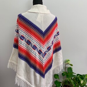 May include: White knitted poncho with a geometric pattern in shades of blue, red, and orange. The poncho has a fringe border.