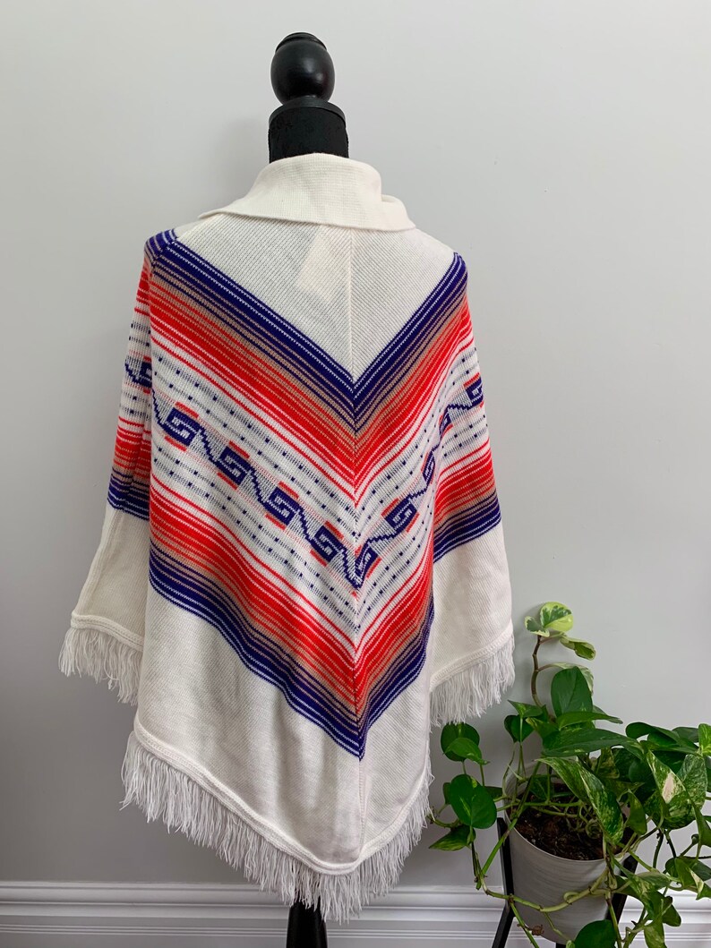 May include: White knitted poncho with a geometric pattern in shades of blue, red, and orange. The poncho has a fringe border.