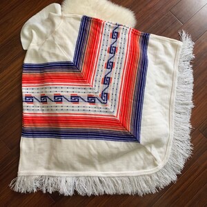 May include: White knitted poncho with a geometric pattern in red, blue, and orange. The poncho has a fringe border.