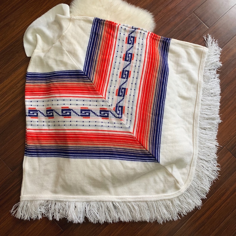 May include: White knitted poncho with a geometric pattern in red, blue, and orange. The poncho has a fringe border.