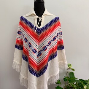 May include: White knitted poncho with a blue, orange, and red geometric pattern. The poncho has a fringe trim and a tie at the neck.