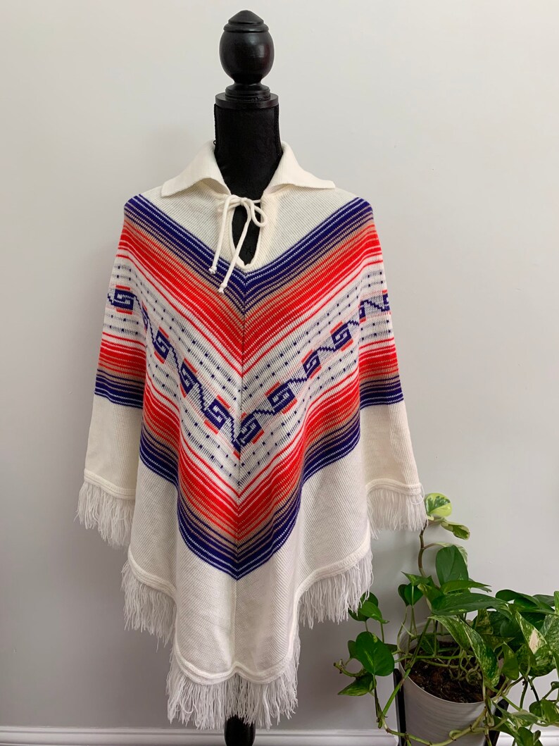 May include: White knitted poncho with a blue, orange, and red geometric pattern. The poncho has a fringe trim and a tie at the neck.