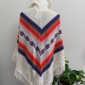 May include: White knitted poncho with a geometric pattern in shades of blue, red, and orange. The poncho has a fringe trim and a high collar.