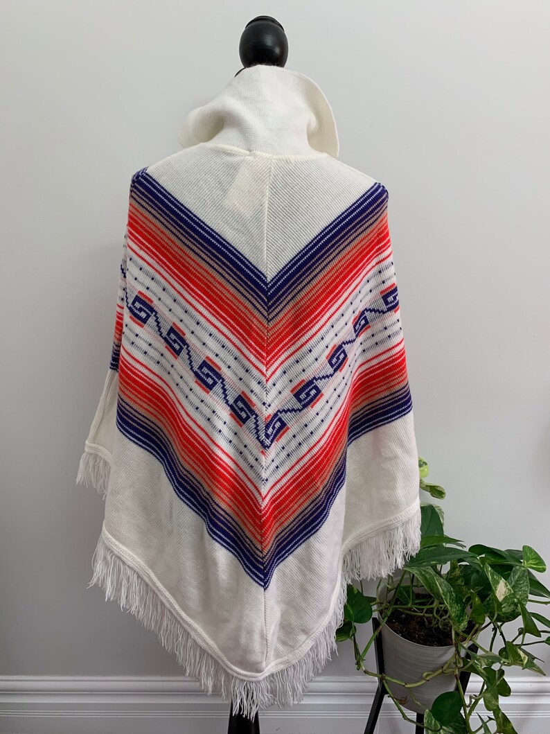 May include: White knitted poncho with a geometric pattern in shades of blue, red, and orange. The poncho has a fringe trim and a high collar.