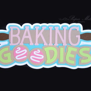 May include: A blue and white paper craft sign with the text "Baking Goodies" in pink, brown, and green letters. The sign features a brown rolling pin and two pink and white cookies.