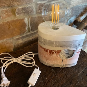 Op de afbeelding: Een witte betonnen tafellamp met een roze en grijs gestreept ontwerp. De lamp heeft een ronde, heldere glazen kap met een gloeilamp met filament. De lamp heeft een wit snoer en een schakelaar.